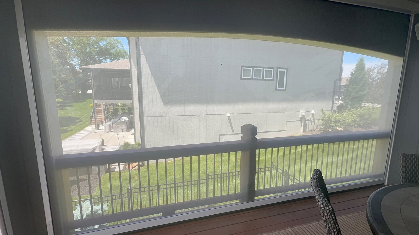 A covered porch with a motorized exterior screen lowered across its wide opening; the deck railing, the lawn and the house next door show through the screen