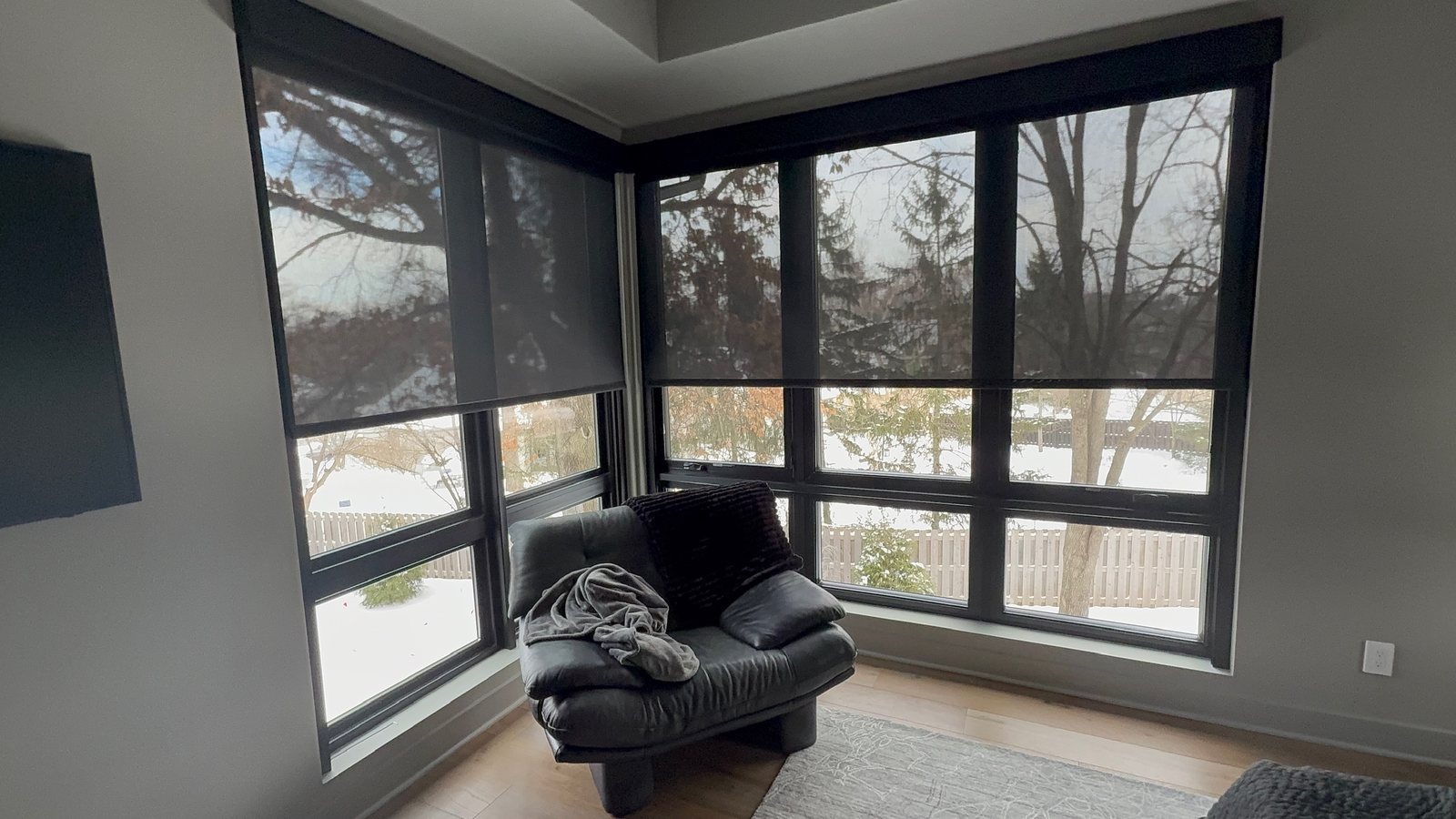 A corner of tall windows in a sitting room, with dark solar shades lowered over the upper panes; the trees and a snowy yard still show through the fabric