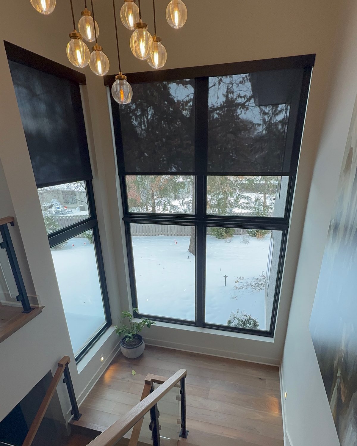 A two-story stairwell with tall windows on two walls under a cluster of glass pendant lights; the narrow window's shade is lowered all the way and the wide window's shade halfway