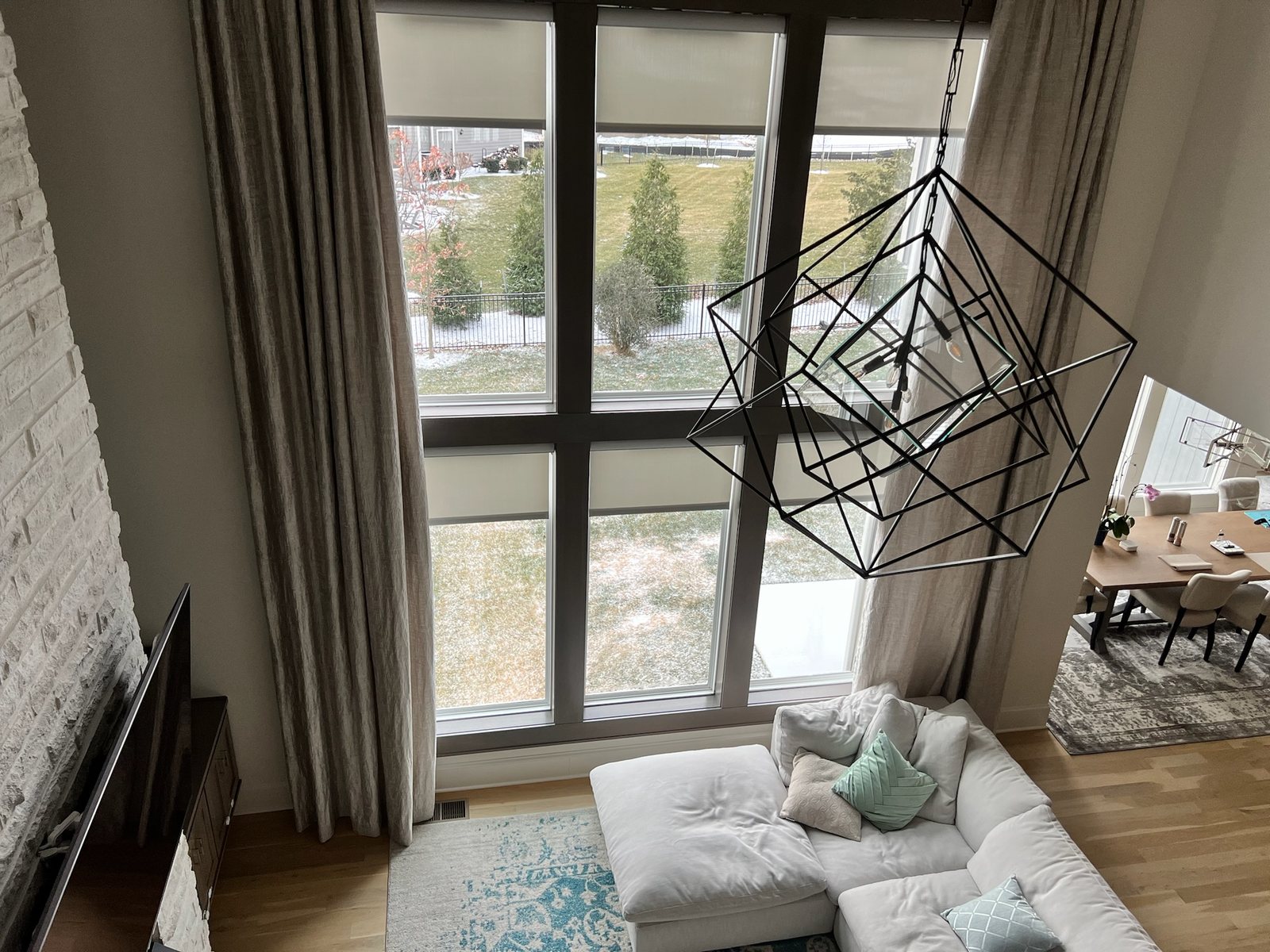A two-story window wall above a family room, with a roller shade lowered partway in each pane of the upper and lower rows, drapery panels at the sides and a geometric pendant in front