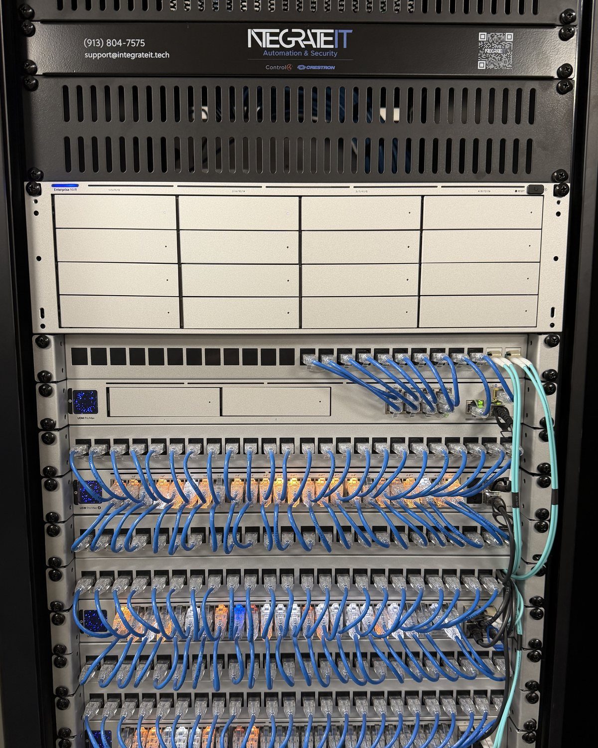 A network rack under an IntegrateIT faceplate: a camera recorder, a firewall and network switches, every port patched with short blue cables