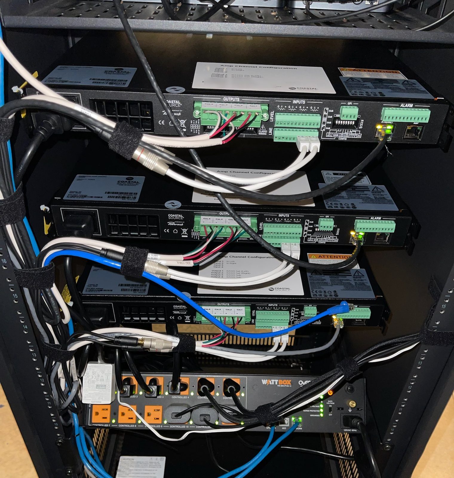 The back of an equipment rack: three amplifiers, each with a printed channel sheet, above a surge-protected power unit, with the cables bundled and strapped